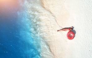 Aerial view of the beautiful young lying woman with pink donut swim ring on the white sandy beach near blue sea with waves at sunset. Summer holiday. Top view of slim girl, clear water. Indian Ocean