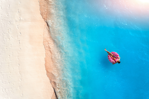 Aerial view of a young woman swimming with the donut swim ring in the clear blue sea with waves at sunset in summer. Tropical aerial landscape with girl, azure water, sandy beach. Top view. Travel