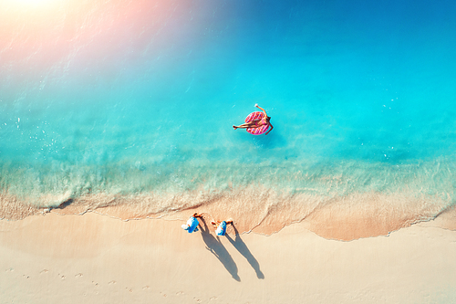 Aerial view of a young woman swimming with the donut swim ring in the blue sea, waves and walking people at sunset in summer. Tropical aerial landscape with girl, azure water, sandy beach. Top view