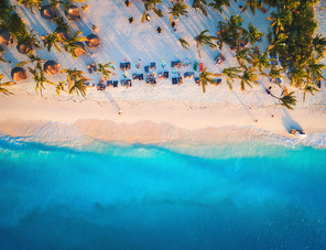 Aerial view of umbrellas, palms on the sandy beach of Indian Ocean at sunset. Summer travel in Zanzibar, Africa. Tropical landscape with palm trees, parasols, people, sand, blue water, waves. Top view