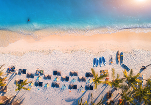 Aerial view of umbrellas, palms on the sandy beach of blue sea at sunset. Summer travel in Zanzibar, Africa. Tropical landscape with palm trees, parasols, people, sand, waves. Top view from the air