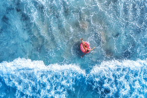 Aerial view of a young woman swimming with the pink donut swim ring in clear blue sea with beautiful waves at sunny day in summer. Tropical aerial landscape with girl, azure water. Top view. Travel