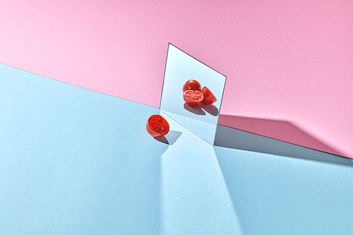 A mirror with whole and halved tomatoes and a ripe half of a vegetable on a double pink-blue paper background with copy space