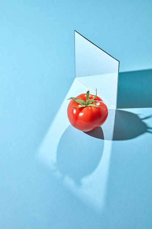 Red tomato with a green stem and a mirror on a blue background with shadows and a copy of a spacer. Creative food composition