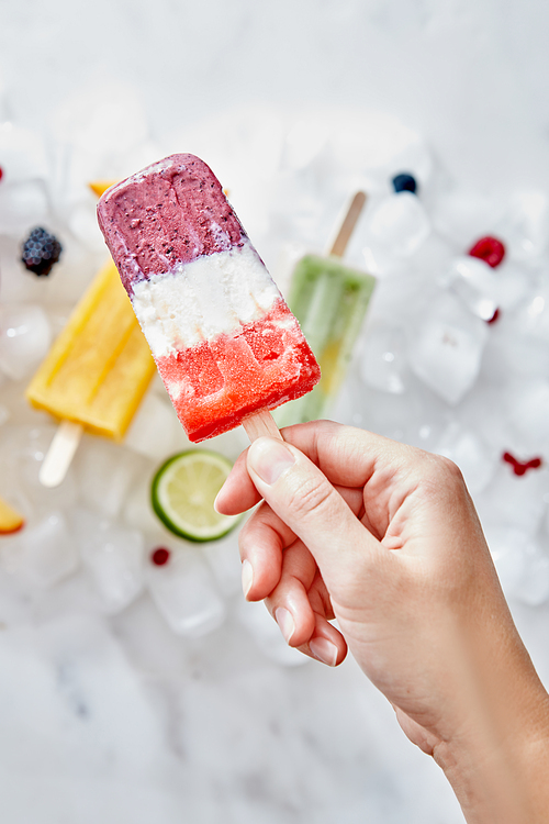 Appetizing fruit berry frozen smoothies lolly is holding the girl's hand against the background of ice cubes with fruits and ice cream. Healthy dessert. Flat lay