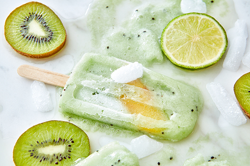 Splashes of Fruit ice cream on a stick and pieces of ice, lime and kiwi on a gray background.Summer dessert lolly with a piece of peach. Flat lay