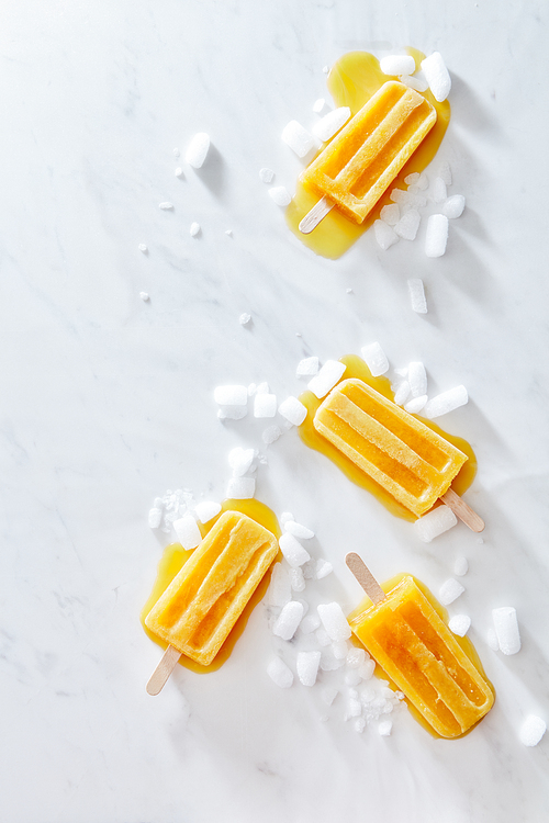 Mango ice cream on a stick with ice slices and splashes on a gray marble table with a copy of the space. Homemade summer dessert. Flat lay