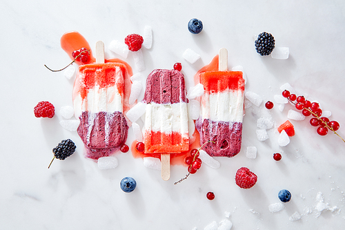 Homemade vegan frozen blood red natural juice popsicles over ice with berries on marble background, top view. Summer mood