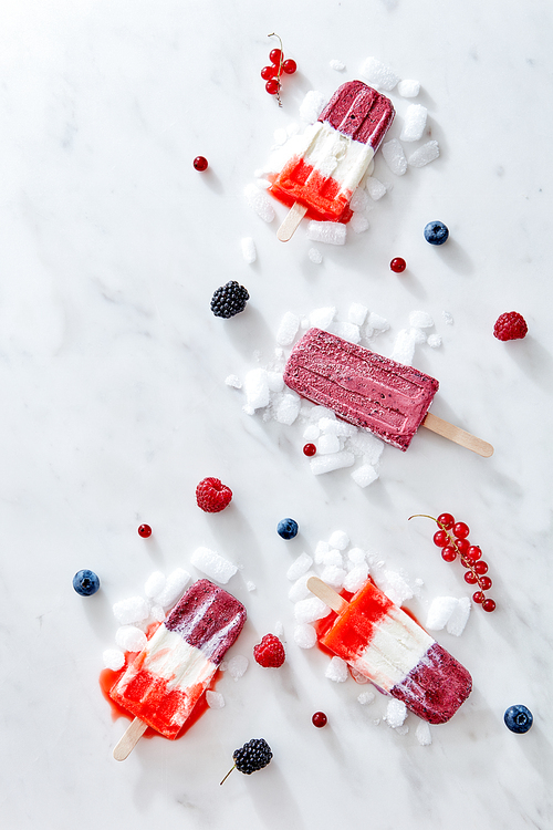 Tricolor strawberry and raspberry ice cream popsicles over ice with berries on marble background, top view and copy space