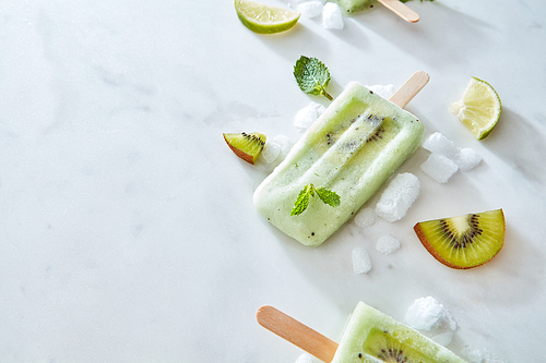 Frozen green smoothies lolly with a slice of kiwi on a stick with mint leaves, ice and pieces of lime and kiwi on a gray marble table with space for text. Flat lay