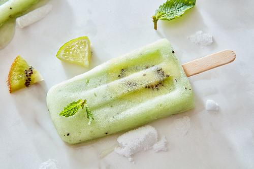 Close-up Frozen fruit juice on a stick with a piece of fresh kiwi on a gray marble background with mint leaves and pieces of lime, ice and kiwi. Copy space for text. Flat lay