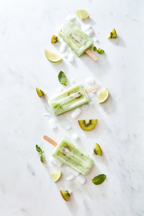Food pattern of lime ice cream lollywith a slice of kiwi, fresh mint leaves with ice slices, lime and kiwi on a gray marble background with space for text. Flat lay