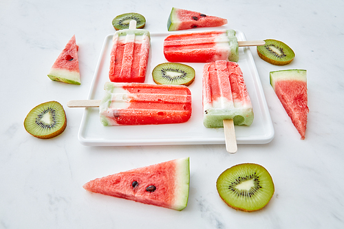 Creative pattern from different pieces of fruit and berry color ice cream on a stick in the shape of a square in a white plate on a gray marble background .