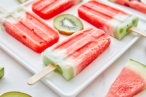 Watermelon ice cream on a stick presented in the form of a square with pieces of kiwi and juicy pieces of watermelon in a plate on a gray marble background. Summer dessert