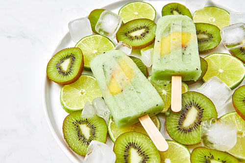 Natural organic frozen juice on a stick with a slice of mango in a bowl with pieces of lime, kiwi and ice cubes on a gray marble table with a copy of the space for text. Top view