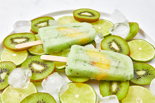 Close-up of healthy home-made fruit ice cream on a stick with a slice of peach in a plate with pieces of lime, kiwi and cold ice cubes. Organic dietary dessert