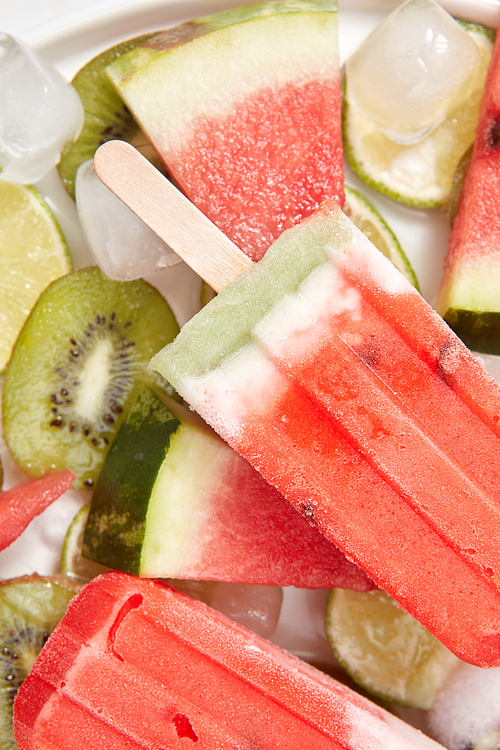 Close-up healthy refreshing homemade ice cream lolly in a white plate with melting ice cubes and pieces of kiwi, lime, watermelon. Flat lay