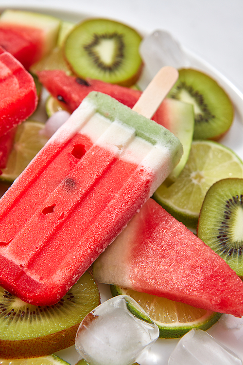 Multicolored berry frozen smoothies lolly with pieces of kiwi, lime, watermelon and ice cubes in a plate on a gray background close-up. Flat lay