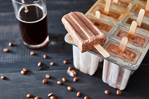 Appetizing coffee ice popsicle on a stick with a glass of cold coffee on a black wooden background with coffee beans. Top view