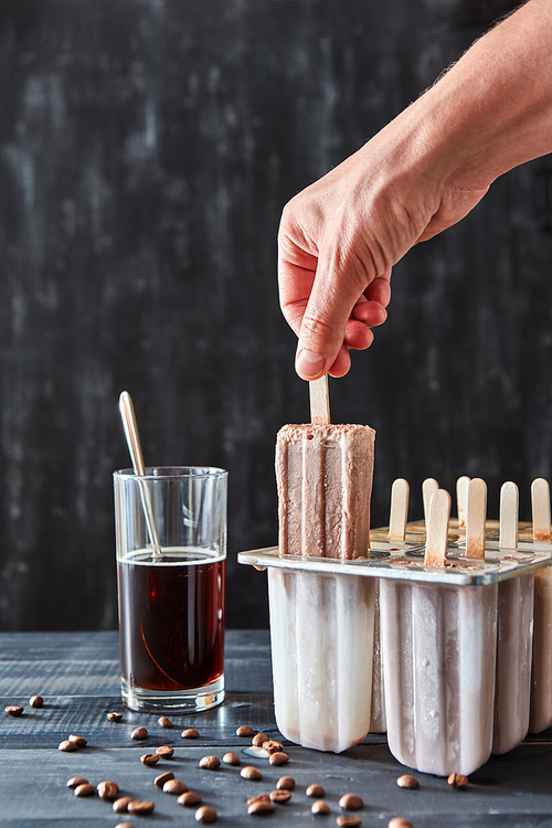 Coffee ice lolly. A woman's hand takes ice cream with plastic molds, a glass of coffee and coffee beans on a black wooden table. Copy of space
