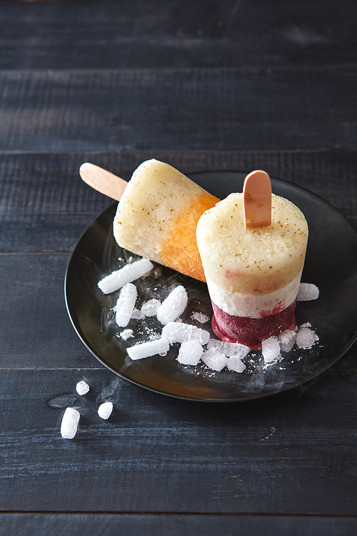 Homemade smoothies lolly on a stick. Multicolored berry, coffee ice cream with ice cubes in a black plate on a dark wooden background with space for text. Flat lay