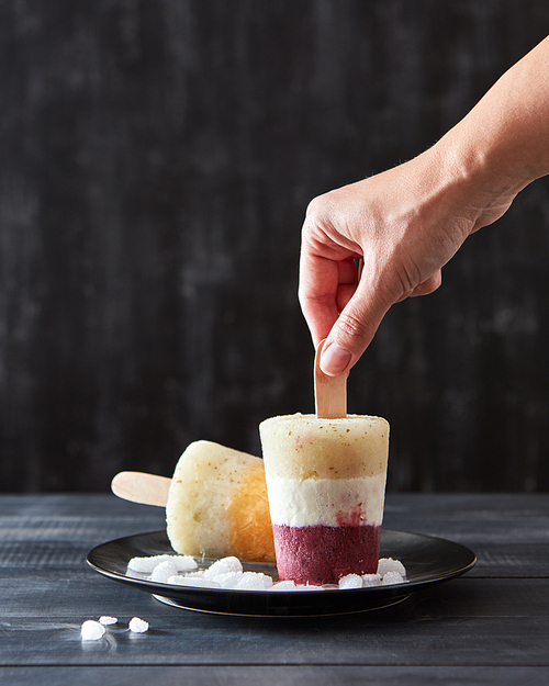 A woman's hand takes a fruity coffee homemade ice cream lolly in a black plate with ice cubes on a dark wooden background with copy space.