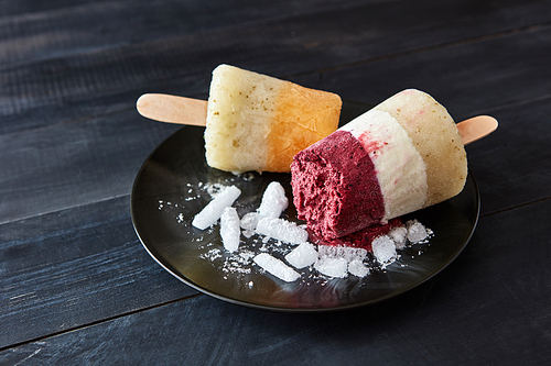 Different coffee, fruit ice cream on a stick are presented in a black plate with slices of ice on a dark wooden background with space for text. Healthy sweet dessert