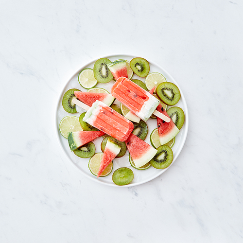 Fruit frozen smoothies on a stick in the form of a piece of watermelon in a plate with ice cubes and pieces of fruit on a gray background with space for text . Ice cream lolly. Flat lay