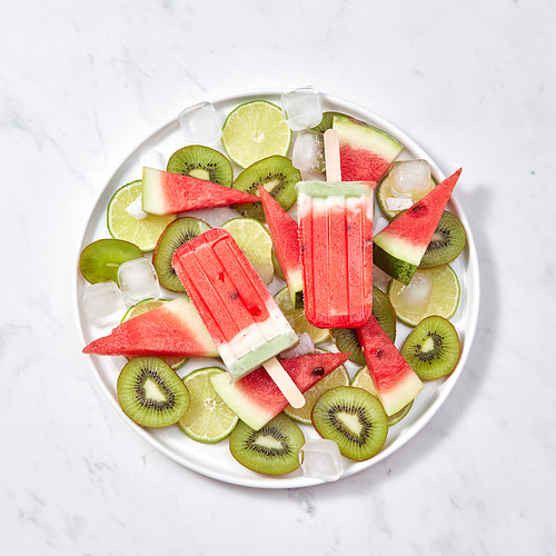 Colorful icy popsicle. Appetizing homemade watermelon ice cream in a plate with slices of lime, watermelon, kiwi and ice cubes on a gray marble table with a copy of the space. Flat lay