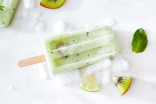 Homemade ice cream on a stick with kiwi and a mint leaf on a white background with ice slices. Flat lay