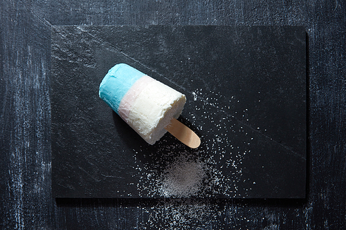 Homemade white blue ice cream lolly decorated with coconut chips on a black slate on a dark concrete background with copy space. Flat lay