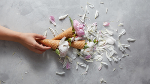 Sweet cones in a woman hand with gentle pink and white peony flowers, petals on a gray concrete background, place for text. Top view. Concept of congratulations for Valentines Day.