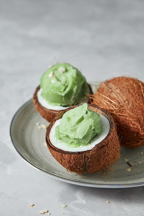 Homemade green ice cream in a coconut shell with whole coconut on a gray concrete background, place for text. Summer dessert.