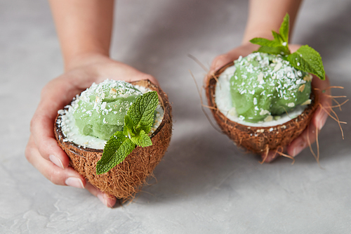 Womans hands take two halves of coconut with homemade green ice cream above a gray concrete background. Summer dessert for vegan