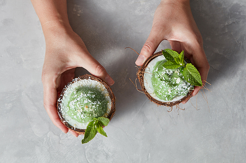A girl hold two halfs of coconut with green sorbet with sprig of mint and coconut chips on a gray concrete. Top view. Summer dessert for vegan