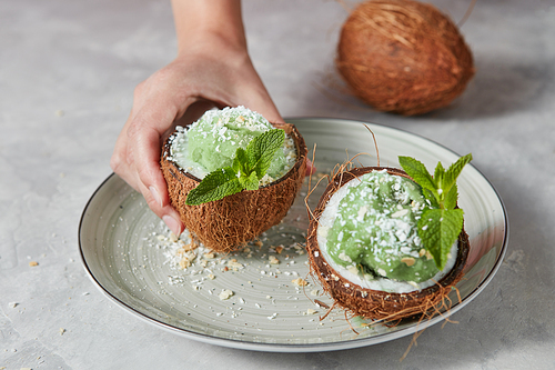 Green mint sorbet in the coconut shell with green leaves holding a girl hand above a plate on a stone background. Summer dessert.