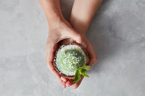Delicious green dessert, ice cream in a coconut shell in a girls hands above a gray concrete background with place for text. Top view.