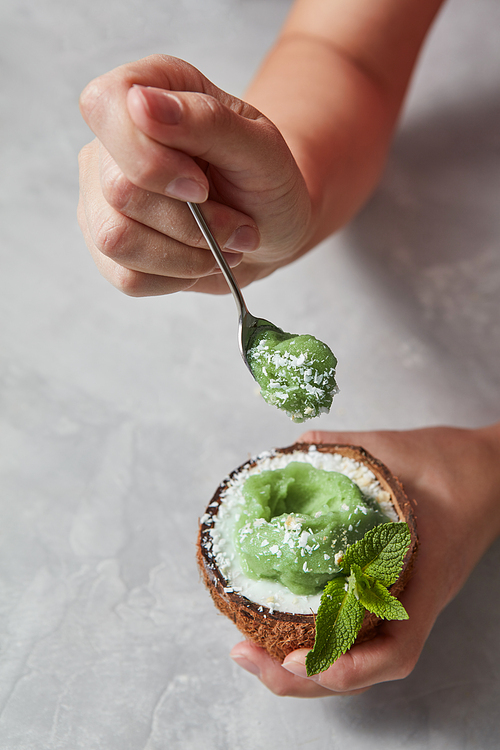 Green mint sorbet ice cream in the coconut shell with green leaves in the hands with spoon above a stone background. Summer dessert for vegan