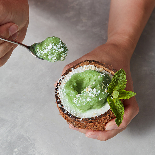 Close-up view a half of coconut with green mint ice cream with sprig of mint and coconut chips in the female hands above a marble gray background. Summer dessert for vegetarian.
