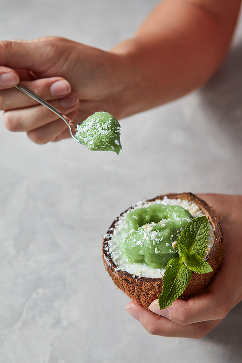 Woman's hands holding organic green sorbet in a coconut with spoon above a gray background with place for text. Summer dessert for vegetarian.