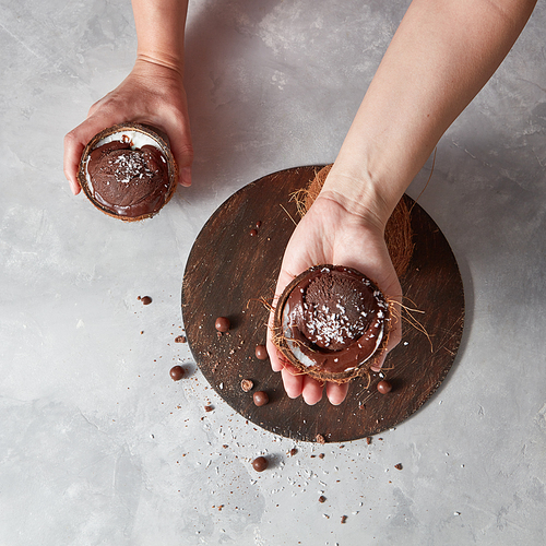 Homemade chocolate dessert in a coconut shell holding womans hands above round board on a gray background with place for text. Summer dessert.