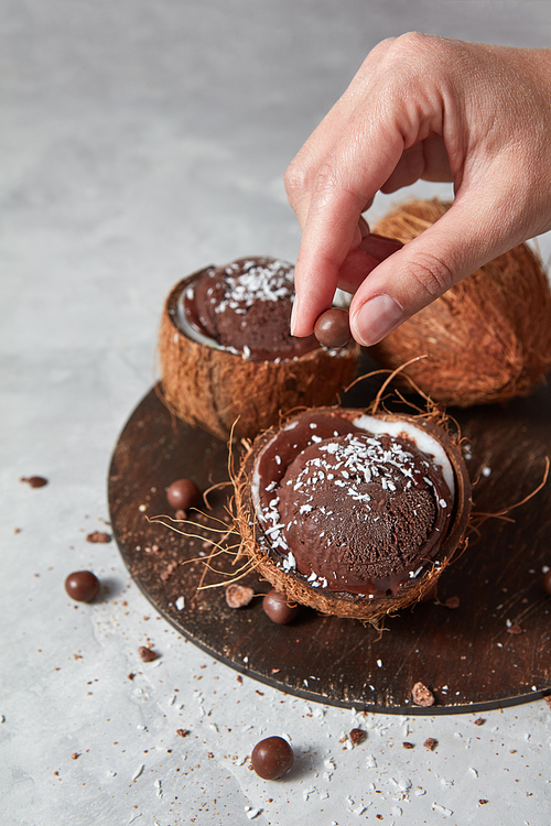 Chocolate brown homemade dessert in a coconut shell with girls hand decorating ice cream on a gray concrete background with place for text. Summer dessert for vegetarian.