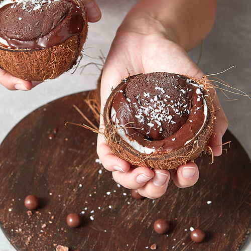 Delicious melted cocolate dessert, ice cream in a coconut shell in a girls hands above a wooden board on a gray background. Vegetarian concept of diet eating