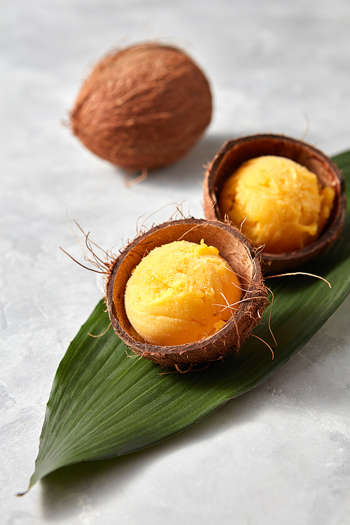 yellow sorbet ice cream in the coconut shell with green palm leaves on stone background. Summer concept