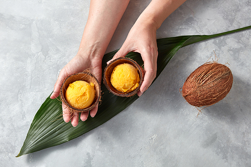 Top view of two halves of a coconut shell with yellow ice cream balls over a green tropical leaf of a plant that hold the woman's hands and a whole coconut on a gray marble table. Summer concept.