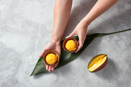 Woman hands hold frozen yellow mango sorbet in the coconut shell with half fresh mango on palm leaves on gray stone background. Flat lay