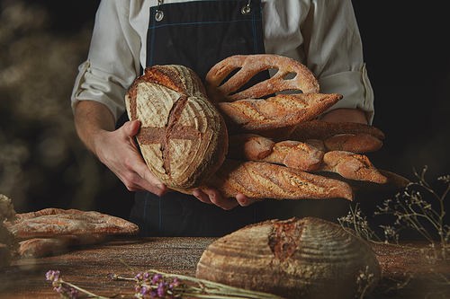 Baker in a black apron holds a variety of bread, rye bread, baguette,fougas, on the background of an old brown table