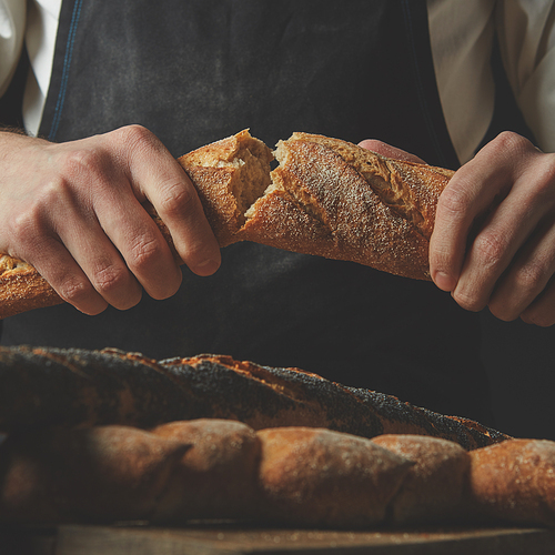 Close-up organic delicious baguette halves in the hands of a man on the background of a black apron and a brown wooden table