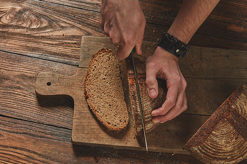 top view of fresh bread cut into a man's hands on a wooden board.in the background of a brown table flat lay