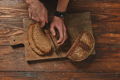 Hands of a young man cut a dark bread on a wooden board against the background of an old wooden table flat lay